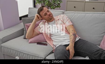 Un uomo rilassato tatuato che si rilassa su un divano in un elegante appartamento, con uno smartphone accanto a lui, incarnando la vita familiare. Foto Stock