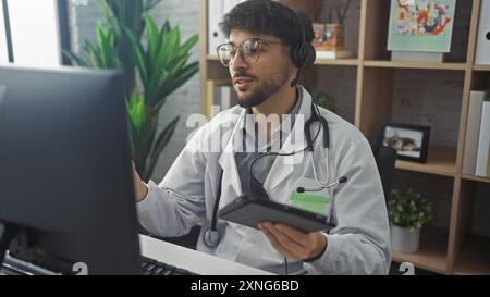 Un giovane uomo barbuto con una cuffia e un cappotto bianco regge un tablet in un ufficio della clinica. Foto Stock