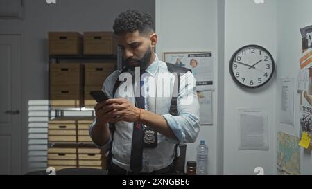 Detective ispanico che analizza gli indizi in un ufficio, si è concentrato sul suo smartphone come potenziale prova con un distintivo visibile. Foto Stock