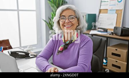 Una donna dai capelli grigi sorridente in ufficio con un laptop, occhiali e sciarpa colorata trasuda professionalità e positività. Foto Stock