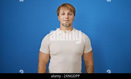 Bel giovane uomo caucasico adulto con barba, capelli biondi e tatuaggi si staglia su uno sfondo blu isolato all'aperto. Foto Stock