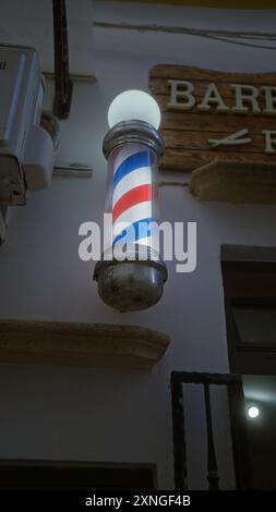 Una vivace asta da barbiere fuori da un barbiere a gallipoli, puglia, italia, illuminata al crepuscolo su un rustico cartello di legno, che incarna il tradizionale groo Foto Stock