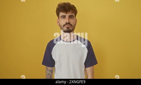 Bellissimo giovane ispanico con barba e tatuaggi in posa su un vibrante sfondo giallo Foto Stock
