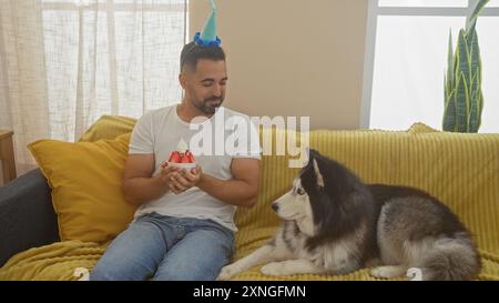 Uomo ispanico che festeggia con il husky siberiano su un divano giallo all'interno, tenendo una torta di compleanno adatta ai cani. Foto Stock