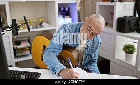 Un anziano barbuto con camicia in denim che scrive a una scrivania con chitarra, microfono e computer in un moderno ufficio domestico. Foto Stock