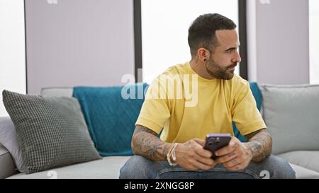 Un uomo ispanico barbuto in camicia gialla si rilassa su un divano, tenendo in mano uno smartphone, in un moderno soggiorno. Foto Stock