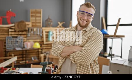 Uomo con barba sicura e le braccia incrociate indossando occhiali protettivi in un'officina di falegnameria Foto Stock