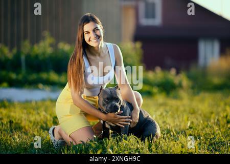 Ritratto all'aperto di una giovane donna bianca con una Bullie americana che si toglie la lingua, una donna europea che coccola il suo cane sullo sfondo di un paesaggio rurale Foto Stock