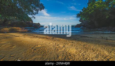 La calda luce del sole bagna un'insenatura rocciosa isolata al tramonto. Le onde calme si infrangono sulla spiaggia sabbiosa, creando un ambiente tranquillo e invitante. Perfetto per gli sfondi, Foto Stock