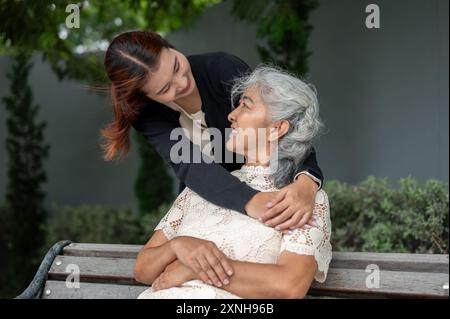 Una bella nipote asiatica abbraccia sua nonna con amore mentre riposa in un giardino insieme, entrambi si guardano l'un l'altro. legame familiare, a. Foto Stock