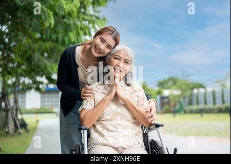 Una bella nipote asiatica sta abbracciando sua nonna, che è su una sedia a rotelle, mentre la cammina in un parco, trascorrendo del tempo insieme, abbracciando con amore Foto Stock