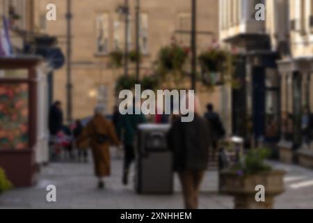 Una vista sfocata delle persone che camminano lungo una strada acciottolata a Bath, Regno Unito, con piante in vaso ed edifici che fiancheggiano il passaggio pedonale. Foto Stock