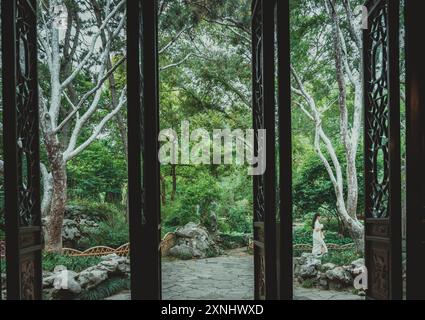 Suzhou, Cina - 11 giugno 2024: Una porta parzialmente aperta in legno rivela una vista di un tradizionale giardino cinese con un sentiero in pietra, lussureggiante vegetazione, un Foto Stock