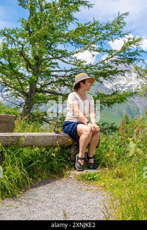 Una donna di 40-45 anni siede circondata da una vegetazione lussureggiante e da una natura colorata. Sullo sfondo, le montagne innevate si innalzano maestosamente sotto il limpido Foto Stock