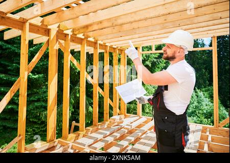 Carpenter costruisce una casa in legno a due piani vicino alla foresta. Uomo barbuto in tuta e casco protettivo che esamina il piano di costruzione. Concetto di moderna costruzione ecologica. Foto Stock