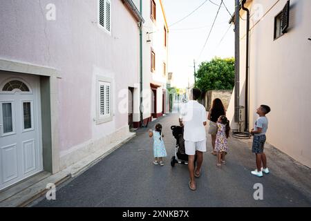 Vista posteriore di una famiglia che cammina lungo una strada stretta con i bambini, mostrando insieme e una rilassante serata estiva a Nin, Croazia. Foto Stock
