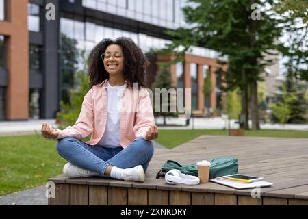 Giovane donna afroamericana che meditava all'aperto, seduta a gambe incrociate su una piattaforma di legno nel parco urbano. Studentessa rilassante in camicia e jeans rosa, con zaino, tazza da caffè e tablet accanto a lei. Foto Stock