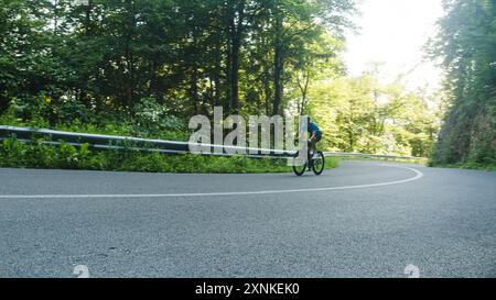 Ciclista maschile da corsa in una maglia blu sportiva, con casco e occhiali, che si sprint su una collina attraverso la foresta. Foto Stock