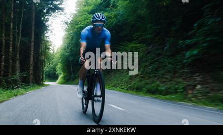 Ciclista maschile da corsa in una maglia blu sportiva, con casco e occhiali, che si sprint su una collina attraverso la foresta. Foto Stock