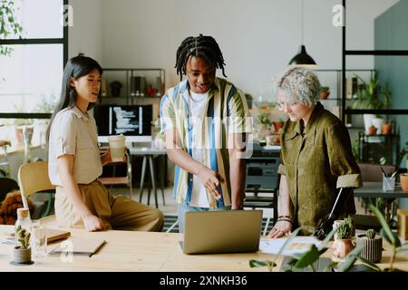 Gruppo di tre diversi giovani manager che indossano abiti casual che guardano lo schermo del notebook mentre lavorano insieme al progetto Foto Stock