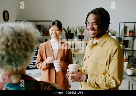 Un gruppo di tre colleghi della generazione Z diversi in piedi in ufficio durante la pausa caffè, ridendo di scherzi divertenti Foto Stock
