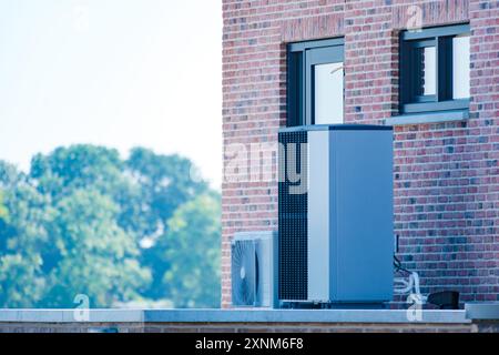 Un'elegante pompa di calore a sorgente d'aria è montata all'esterno di una struttura in mattoni contemporanea, circondata da vegetazione lussureggiante sotto un cielo limpido. pompa di calore della fonte d'aria di traduzione in pompa di calore Foto Stock