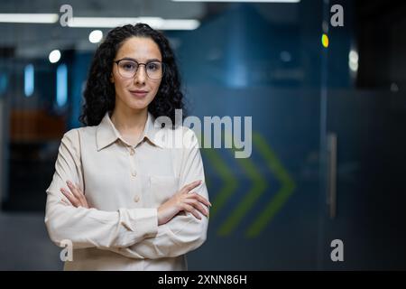 Donna d'affari sicura con capelli ricci e occhiali in piedi con le braccia incrociate in un moderno ambiente da ufficio. L'abbigliamento professionale e l'espressione seria trasmettono leadership e determinazione. Foto Stock