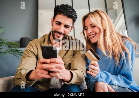 Una giovane coppia che ordina il cibo online utilizzando un'app di consegna su un telefono cellulare. Uomo e donna sono seduti sul divano a casa loro, pagano online con una carta di credito e un cellulare per un viaggio. Foto di alta qualità Foto Stock