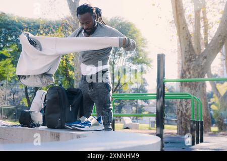 Giovane uomo africano in piedi nel parco la mattina, si sta cambiando i vestiti prima di allenarsi, si toglie il maglione per metterlo nello zaino. Con Foto Stock