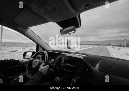 Vista in bianco e nero dall'interno di un'auto di una strada prospettica nel mezzo del deserto nella regione di Zaida in Marocco. Le mani di un anonimo dri Foto Stock