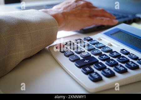 Mani di una consulente donna anziana che immette i numeri calcolati su una calcolatrice in un computer nel suo ufficio Foto Stock