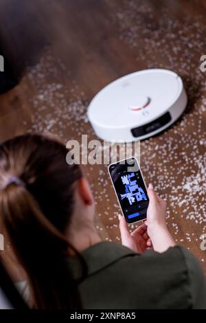 Giovane donna che usa lo smartphone per controllare l'aspirapolvere robot intelligente Foto Stock