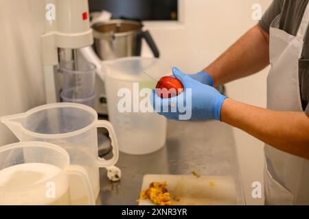 Una persona che indossa guanti blu sta sbucciando una mela rossa in un ambiente di cucina. Sullo sfondo sono visibili varie attrezzature e contenitori per la cucina Foto Stock