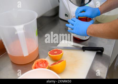 Una persona che indossa guanti blu sta spremendo il succo di pompelmo in un contenitore. I pompelmi a fette si trovano su un tagliere con un coltello. Una caraffa di juic Foto Stock