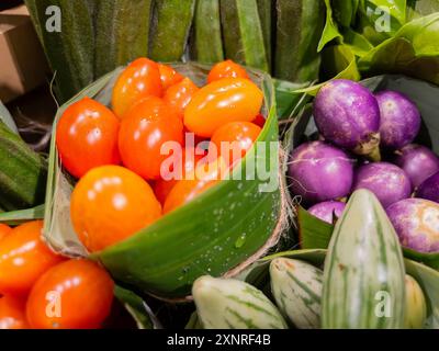 Primo piano di pomodori freschi e varie melanzane, tra cui varietà viola e a strisce, esposte in foglie di banana in un mercato. Foto Stock