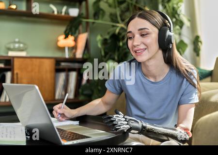 Una giovane donna con protesi e cuffie sembra un libro da lavoro e un notebook Foto Stock