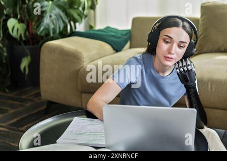 Una giovane donna con protesi e cuffie sembra un libro da lavoro e un notebook Foto Stock