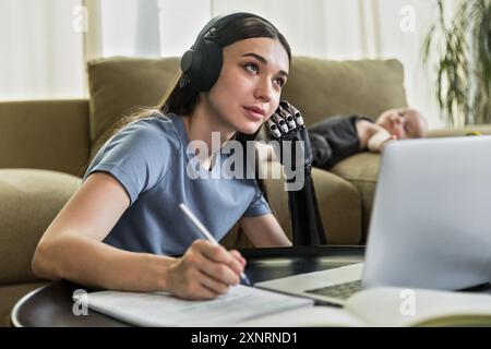 Una giovane donna con protesi e cuffie sembra un libro da lavoro e un notebook Foto Stock