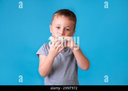 Bambino carino con capelli biondi e lentiggini che mangia una ciambella glassata. Bambini e cibo spazzatura zuccherata. Ragazzo che tiene in mano ciambelle colorate, mangia spazzatura Foto Stock