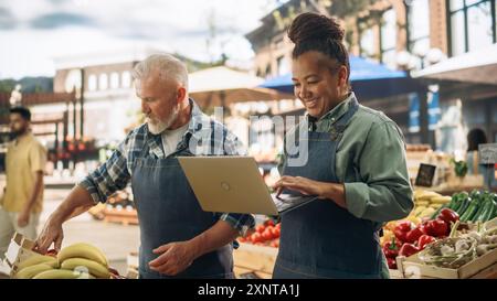 Partner commerciali di sesso femminile e maschile Gestione di un mercato di successo per agricoltori biologici. Coppia di adulti multietnici che parla dell'inventario dei prodotti agricoli durante l'utilizzo di un computer portatile Foto Stock