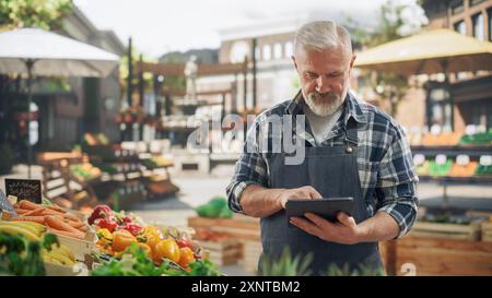 Ritratto di un agricoltore adulto che utilizza un tablet al lavoro mentre vende prodotti agricoli biologici freschi da una situazione di mercato. Uomo di mezza età che gestisce gli ordini online su un sito Web di e-commerce Foto Stock