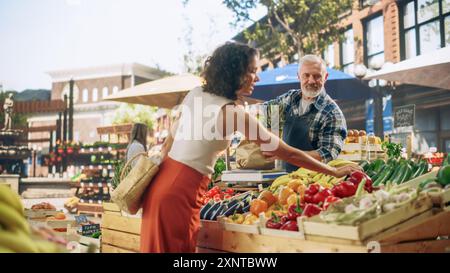 Cheerful Street Vendor gestisce una piccola azienda agricola, vendendo frutta e verdura sostenibili. Uomo di mezza età felice che riempie un sacchetto della spesa di carta riciclata con cibo naturale locale Foto Stock