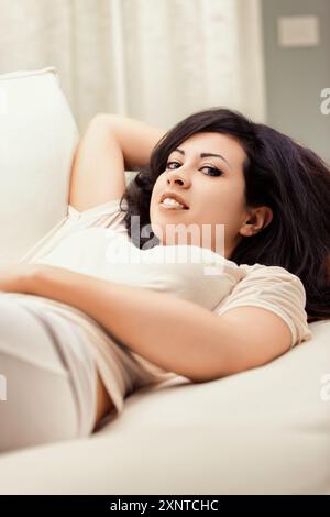 La giovane donna si rilassa sul suo comodo divano a casa, godendosi un momento di tranquillità in un soggiorno elegante e confortevole pieno di luce naturale Foto Stock