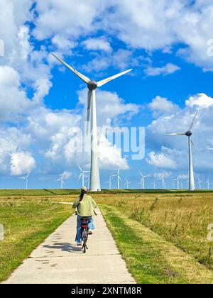 Un ciclista percorre un sentiero circondato da torreggianti turbine eoliche, sfruttando l'energia rinnovabile sotto un cielo blu pieno di nuvole morbide nei Paesi Bassi. Urk Flevoland Foto Stock