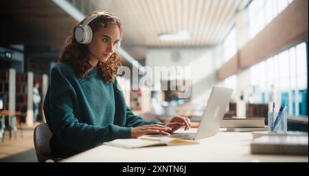 Giovane donna concentrata che studia nella moderna biblioteca, indossa le cuffie e utilizza un laptop. Studentessa impegnata nella ricerca accademica, circondata da libri e interni luminosi Foto Stock