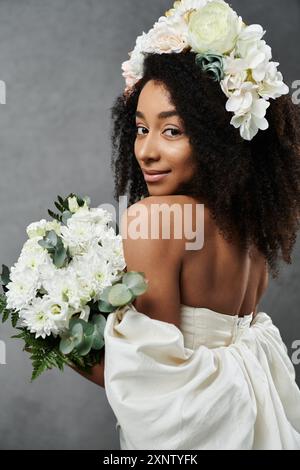 Una sposa afroamericana in abito bianco e corona floreale, con bouquet, posa su sfondo grigio. Foto Stock
