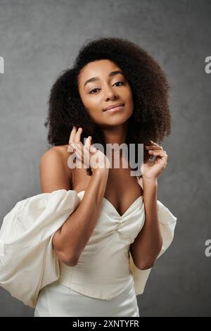 Una sposa afroamericana in un abito da sposa bianco si posa su uno sfondo grigio, irradiando eleganza e gioia. Foto Stock