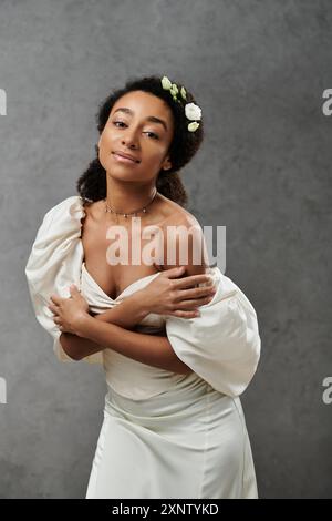 Una splendida sposa afroamericana in un abito da sposa bianco, con fiori nei capelli, posa su uno sfondo grigio. Foto Stock