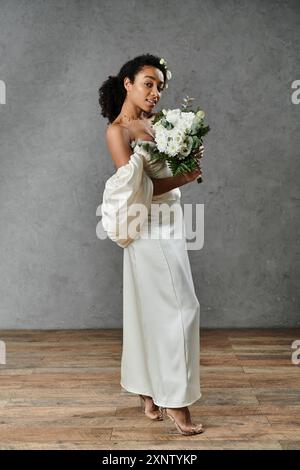 Una splendida sposa afroamericana si erge in un abito da sposa bianco e contiene un bouquet di fiori bianchi. Foto Stock