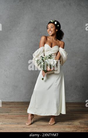 Una sposa afroamericana in un abito da sposa bianco, con fiori nei capelli, posa graziosamente su uno sfondo grigio. Foto Stock